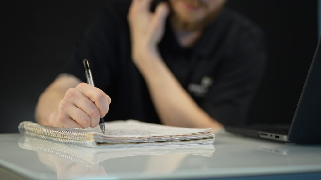 A portrait of a David Williams Electric team member writing a note on a pad of paper with a phone in his opposite hand, while sitting down at a modern white desk in front of a black background.